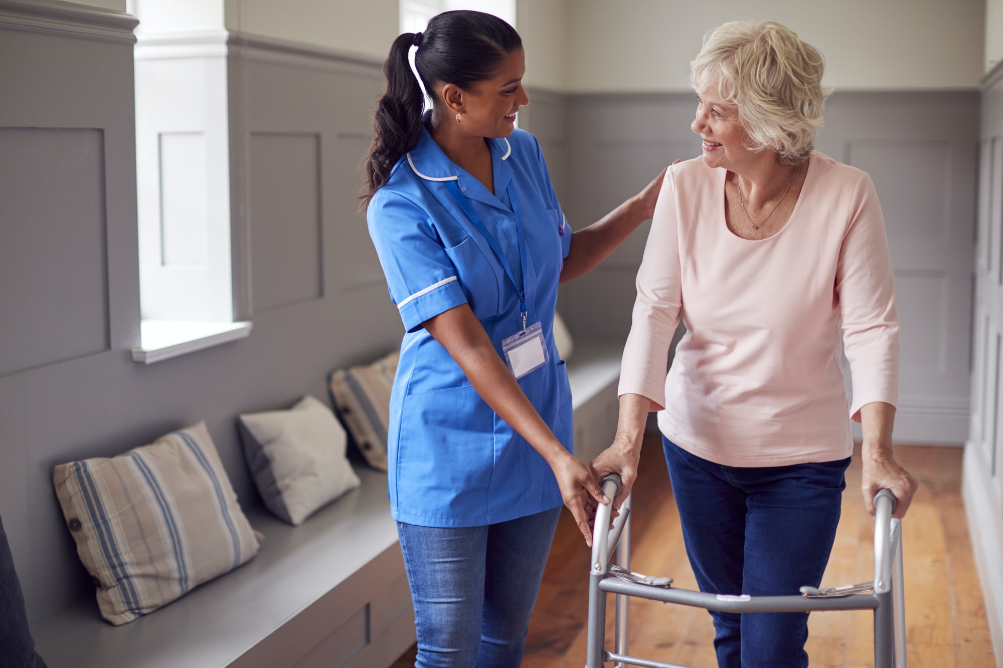Senior Woman At Home Using Walking Frame Being Helped By Female Care Worker In Uniform